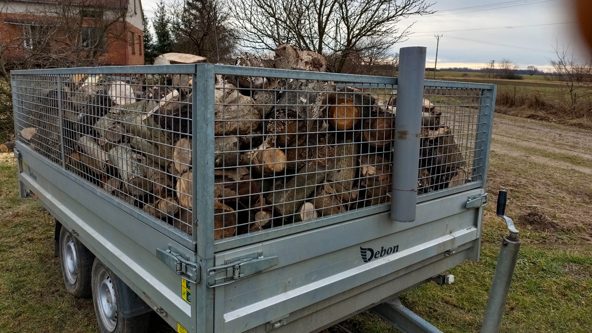 Przyczepa marki Debon wypełniona pociętymi kawałkami drewna, widoczna na tle pola i drzew bez liści pod pochmurnym niebem.