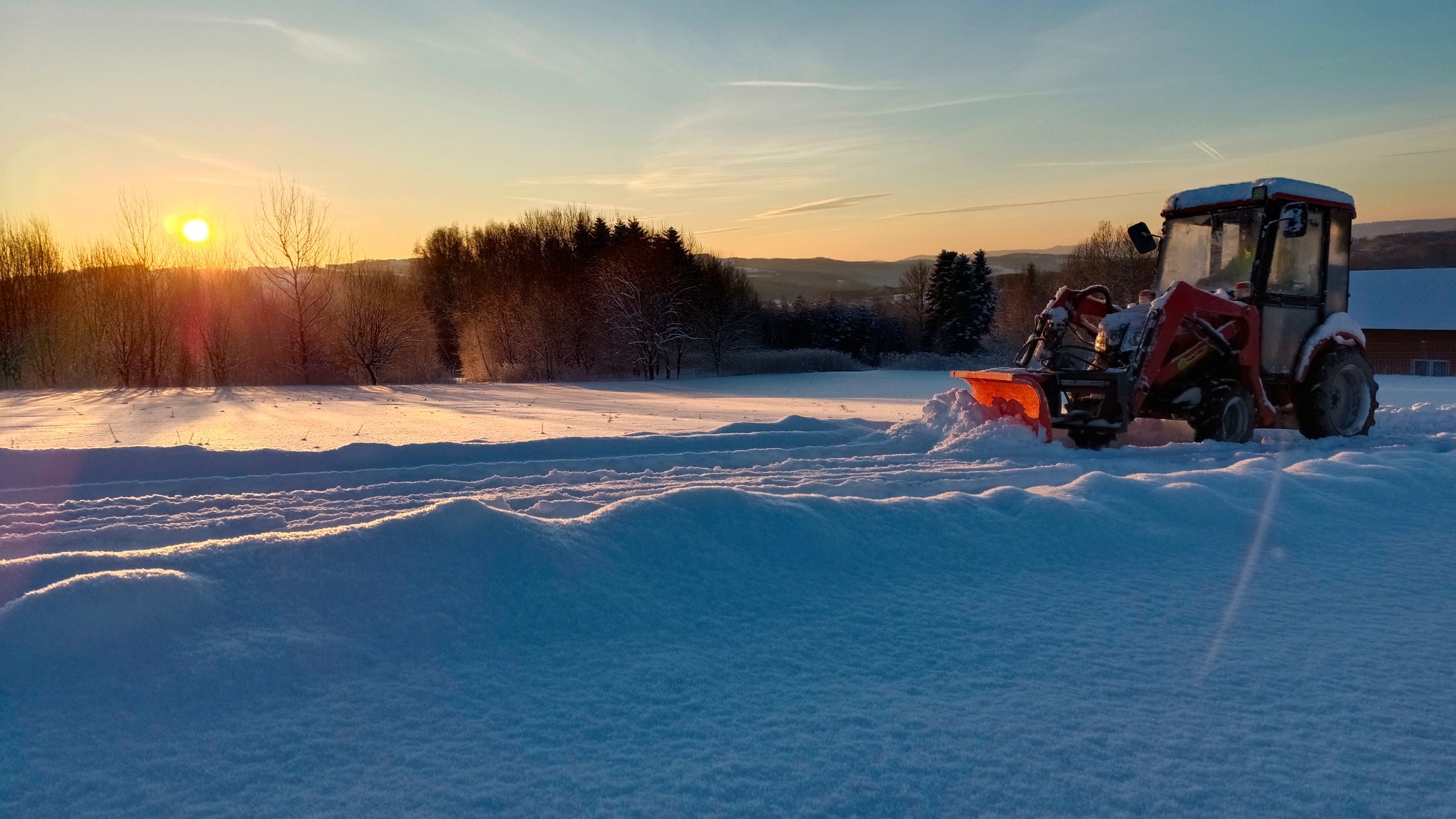 Czerwony traktor z pługiem wirnikowym odśnieża pole pokryte śniegiem podczas wschodu słońca, widoczne ślady opon i wyrzuconego śniegu.