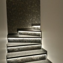 POZIOM remonty i wykończenia - Schody z szarego kamienia z podświetleniem LED, zamontowane na ścianie z dekoracyjną tapetą w odcieniach szarości i złota, oświetlenie punktowe na górze.