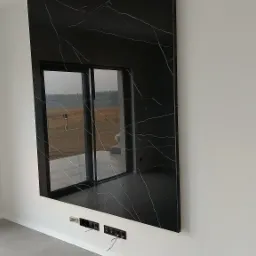 Czarny panel ścienny z wzorem marmuru, zamontowany w nowoczesnym salonie z widocznymi gniazdkami elektrycznymi poniżej i odbiciem krajobrazu w polerowanej powierzchni.