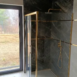 Nowoczesna kabina prysznicowa z czarnymi marmurowymi płytkami i złotymi profilami, widok z boku na przesuwne drzwi i odpływ liniowy w posadzce, z oknem w tle.