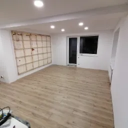 Wnętrze w Charzykowy w trakcie prac: widoczna drewniana konstrukcja pod przyszłą ścianę z płyty karton gips, jasne panele podłogowe i białe ściany z oświetleniem punktowym.