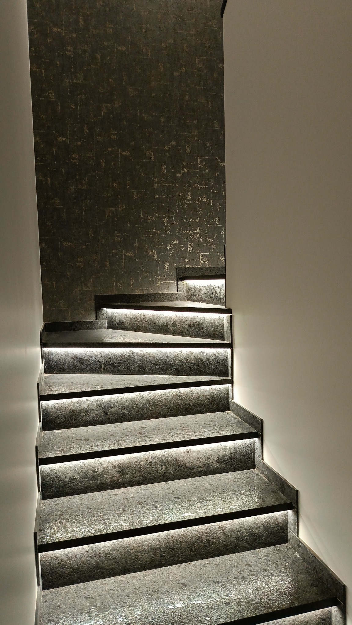 Schody z szarego kamienia z podświetleniem LED, zamontowane na ścianie z dekoracyjną tapetą w odcieniach szarości i złota, oświetlenie punktowe na górze.
