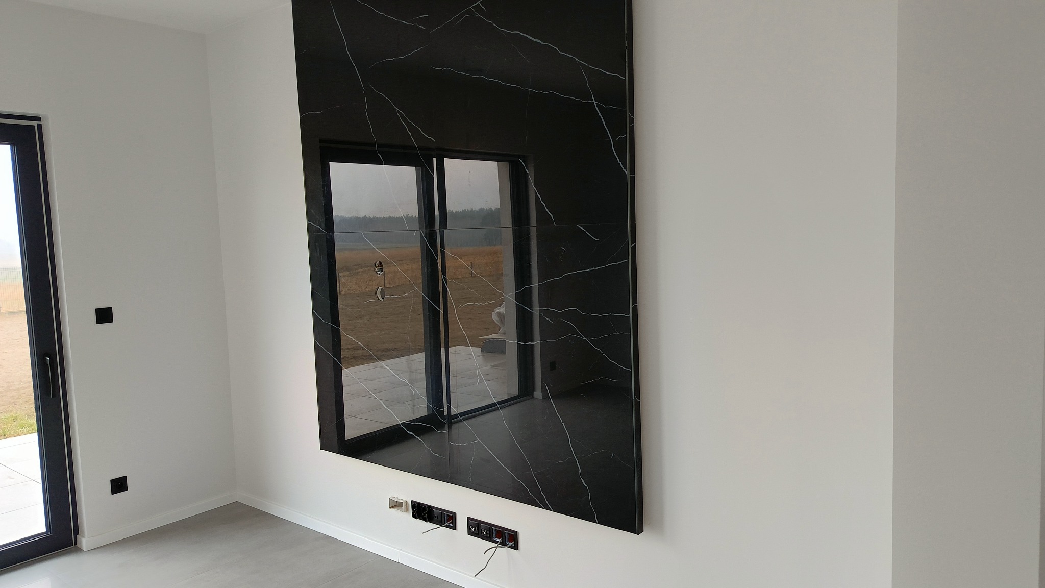 Czarny panel ścienny z wzorem marmuru, zamontowany w nowoczesnym salonie z widocznymi gniazdkami elektrycznymi poniżej i odbiciem krajobrazu w polerowanej powierzchni.