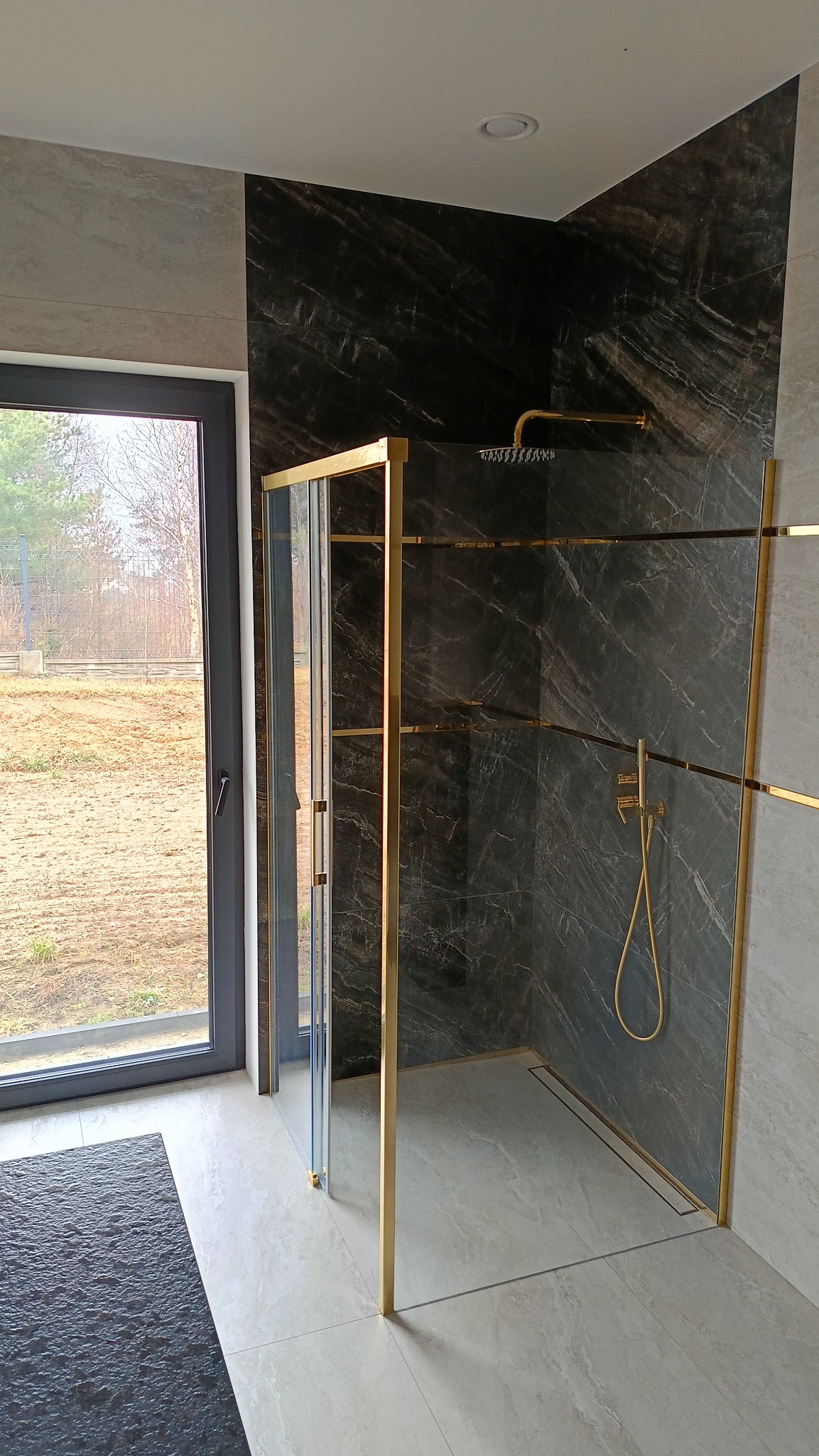 Nowoczesna kabina prysznicowa z czarnymi marmurowymi płytkami i złotymi profilami, widok z boku na przesuwne drzwi i odpływ liniowy w posadzce, z oknem w tle.