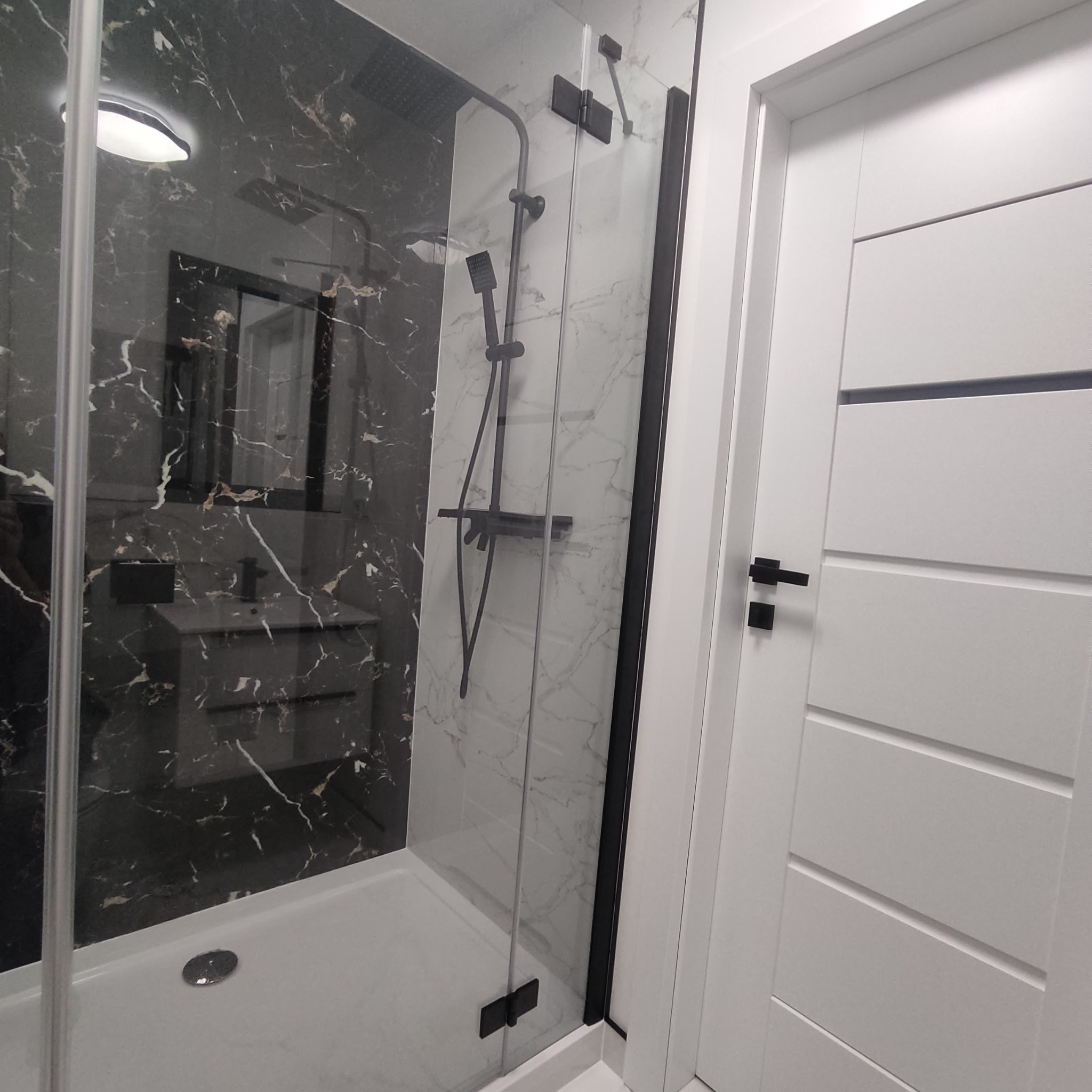 Nowoczesna kabina prysznicowa z czarnymi elementami na tle marmurowej ściany i biała, minimalistyczna drzwi. Widoczna armatura prysznicowa w kolorze czarnym.