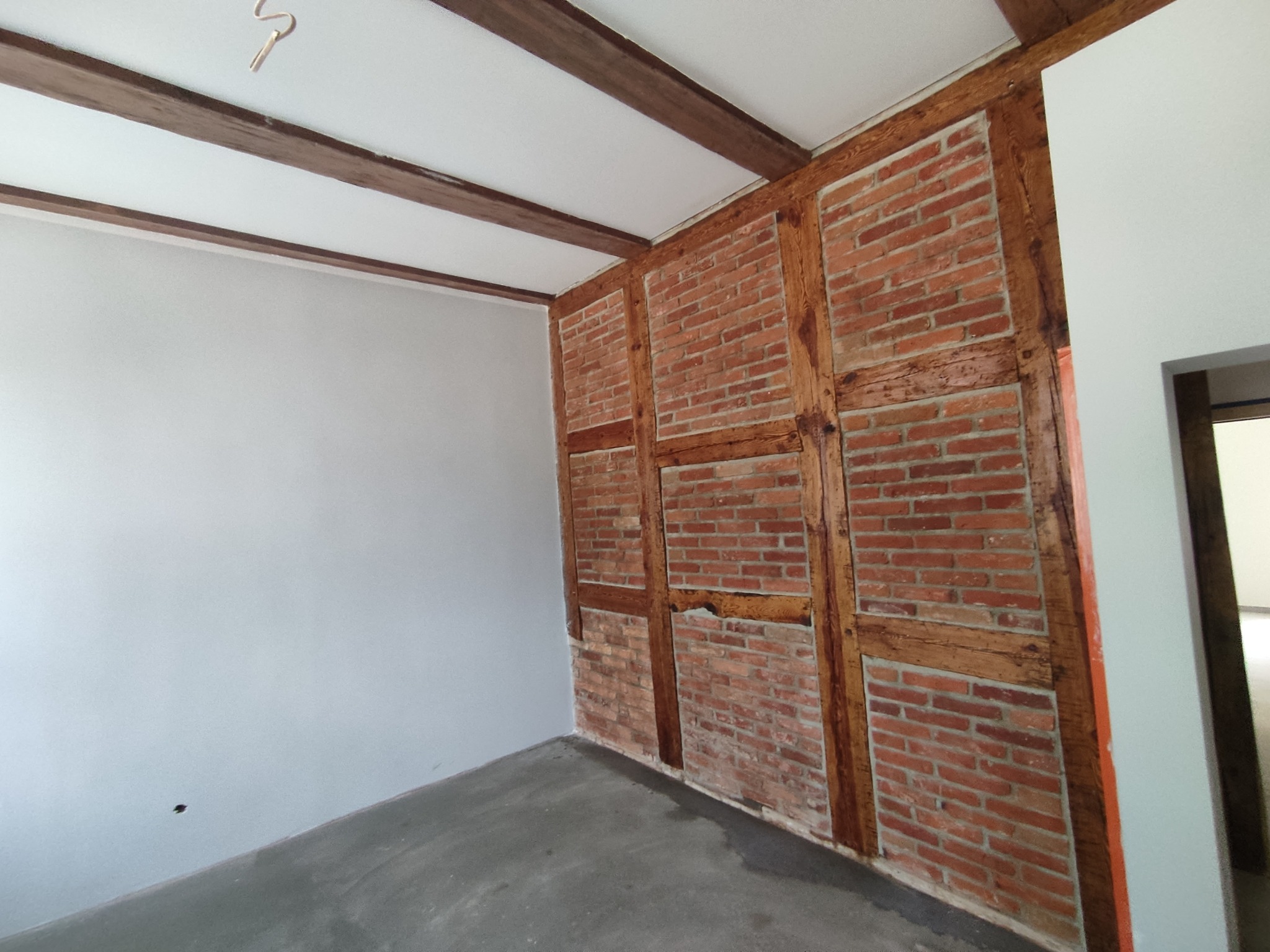 Surowe wnętrze w trakcie prac: ściana z cegły i drewnianych belek, sufit z drewnianymi belkami, szara betonowa podłoga i świeżo pomalowana biała ściana, widoczne niedoskonałości i brak wykończenia.