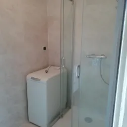 Nowoczesna łazienka z kabiną prysznicową z przesuwnymi drzwiami i pralką automatyczną w rogu, wykończona jasnymi płytkami imitującymi marmur.