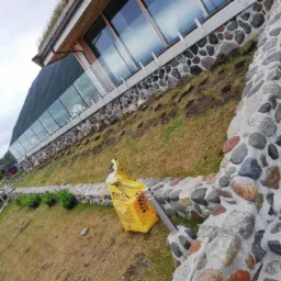 Wyrównywanie terenu pod trawnik w Izabelin-Dziekanówek, widoczny budynek z kamienną elewacją i zielonym dachem, żółty worek z podłożem i wiadro z farbą.