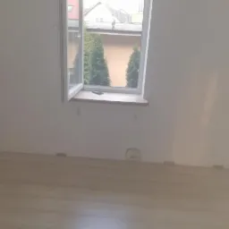 Jasne panele podłogowe w pokoju w trakcie montażu, widoczne kliny dystansowe przy ścianach. Okno z widokiem na zieleń na zewnątrz.