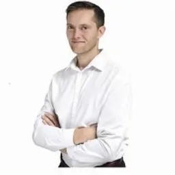 Mężczyzna w białej koszuli z podwiniętymi rękawami, z założonymi rękami na piersi, uśmiecha się patrząc wprost.