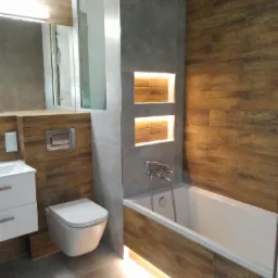 Nowoczesna łazienka z drewnopodobnymi płytkami, podświetlana wanna, wisząca toaleta i szare płytki na ścianie z wnękami oświetlonymi ciepłym światłem.