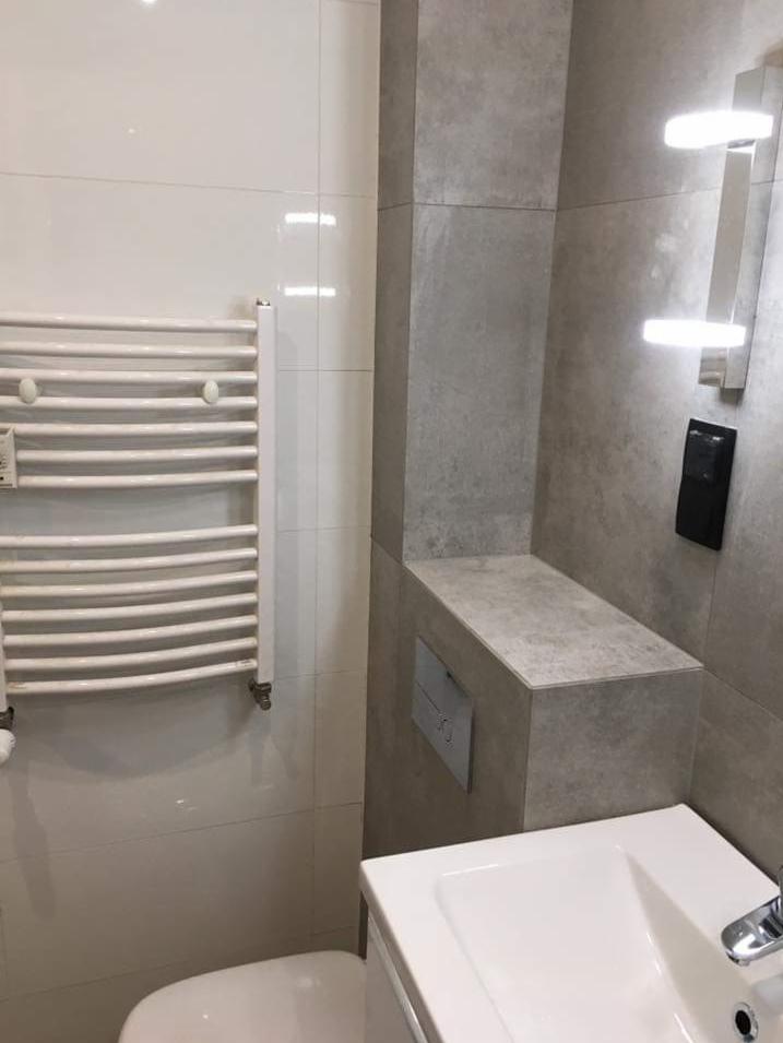 Nowoczesna łazienka z białymi płytkami, grzejnikiem drabinkowym, zabudowaną spłuczką WC i minimalistycznym oświetleniem LED.