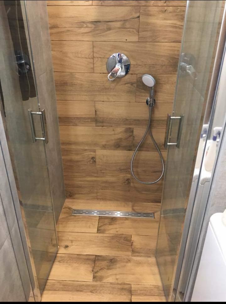 Kabina prysznicowa z odpływem liniowym i baterią natryskową, wykończona płytkami imitującymi drewno, widok z zewnątrz.