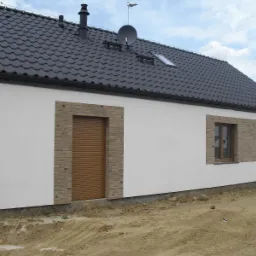 Nowo wybudowany dom jednorodzinny z roletą zewnętrzną w kolorze brązowym, obudowaną dekoracyjną cegłą, oraz oknami z brązowymi ramami, widoczne niedokończone prace ziemne.