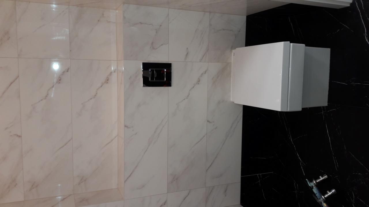 Białe i czarne marmurowe płytki ścienne w łazience z zabudowaną spłuczką i widocznymi rurami.