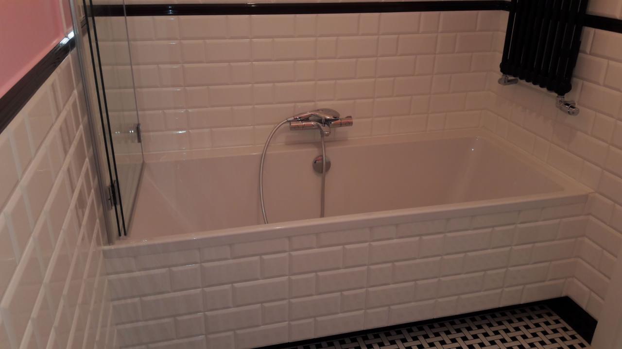 Biała wanna obudowana kafelkami typu metro, czarny grzejnik drabinkowy na ścianie z białych płytek, szklany parawan nawannowy, mozaika czarno-biała na podłodze.