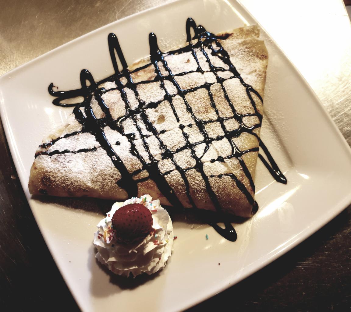 Kremowy naleśnik posypany cukrem pudrem i polany sosem czekoladowym, podany z bitą śmietaną i truskawką na kwadratowym talerzu.