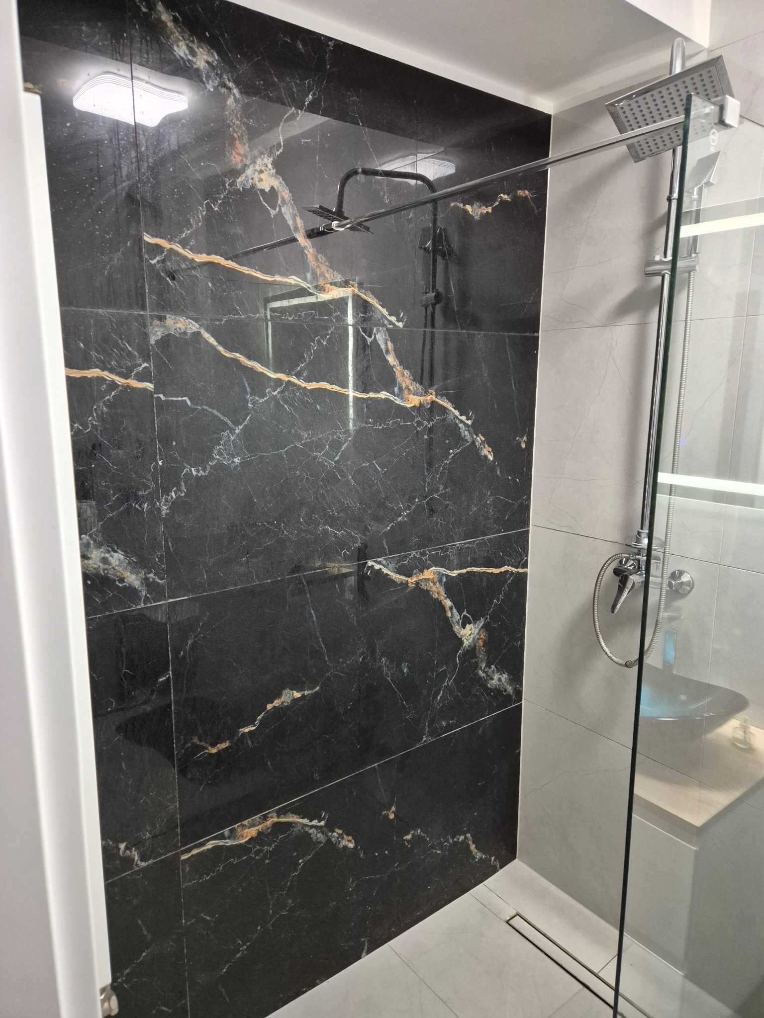 Kabina prysznicowa z czarnymi płytkami imitującymi marmur z złotymi żyłami, szklana ściana, chromowana armatura i odpływ liniowy w jasnych płytkach podłogowych.