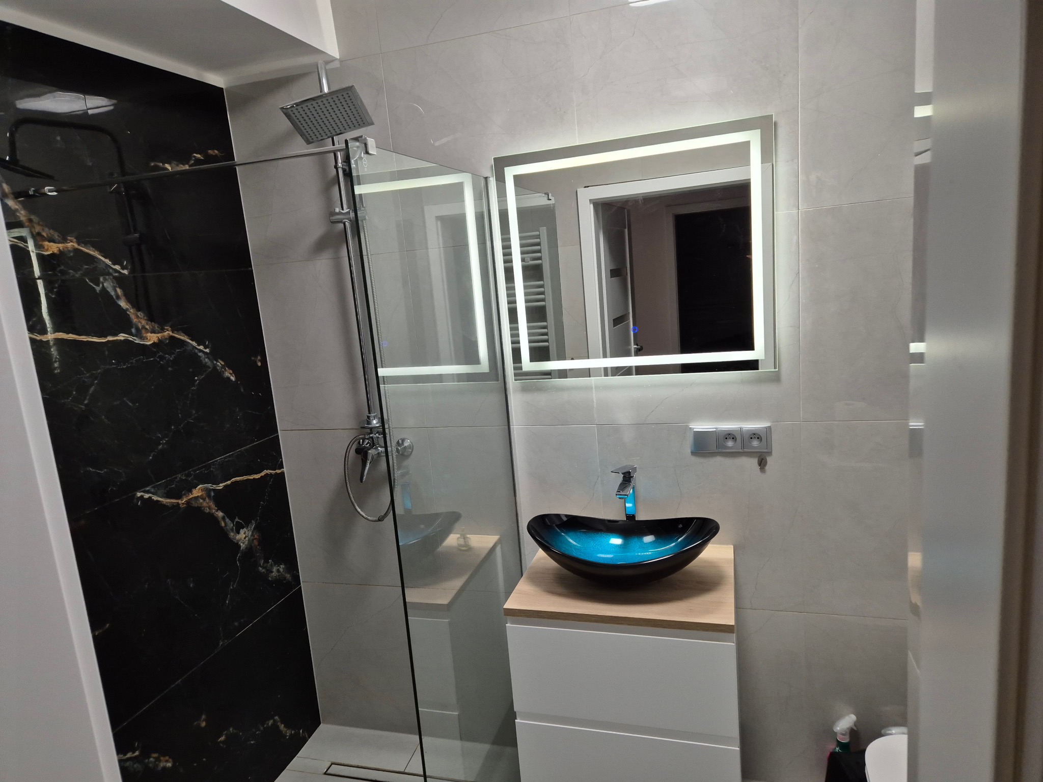 Nowoczesna łazienka z kabiną prysznicową typu walk-in, umywalką w odcieniach turkusu i czerni oraz podświetlanym lustrem. Ściany wyłożone płytkami imitującymi marmur.