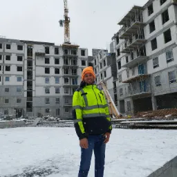 Uśmiechnięty kierownik budowy w pomarańczowej czapce i jaskrawej kamizelce odblaskowej, na tle budowanego bloku z białego pustaka w zimowej scenerii, z widocznym żurawiem budowlanym.