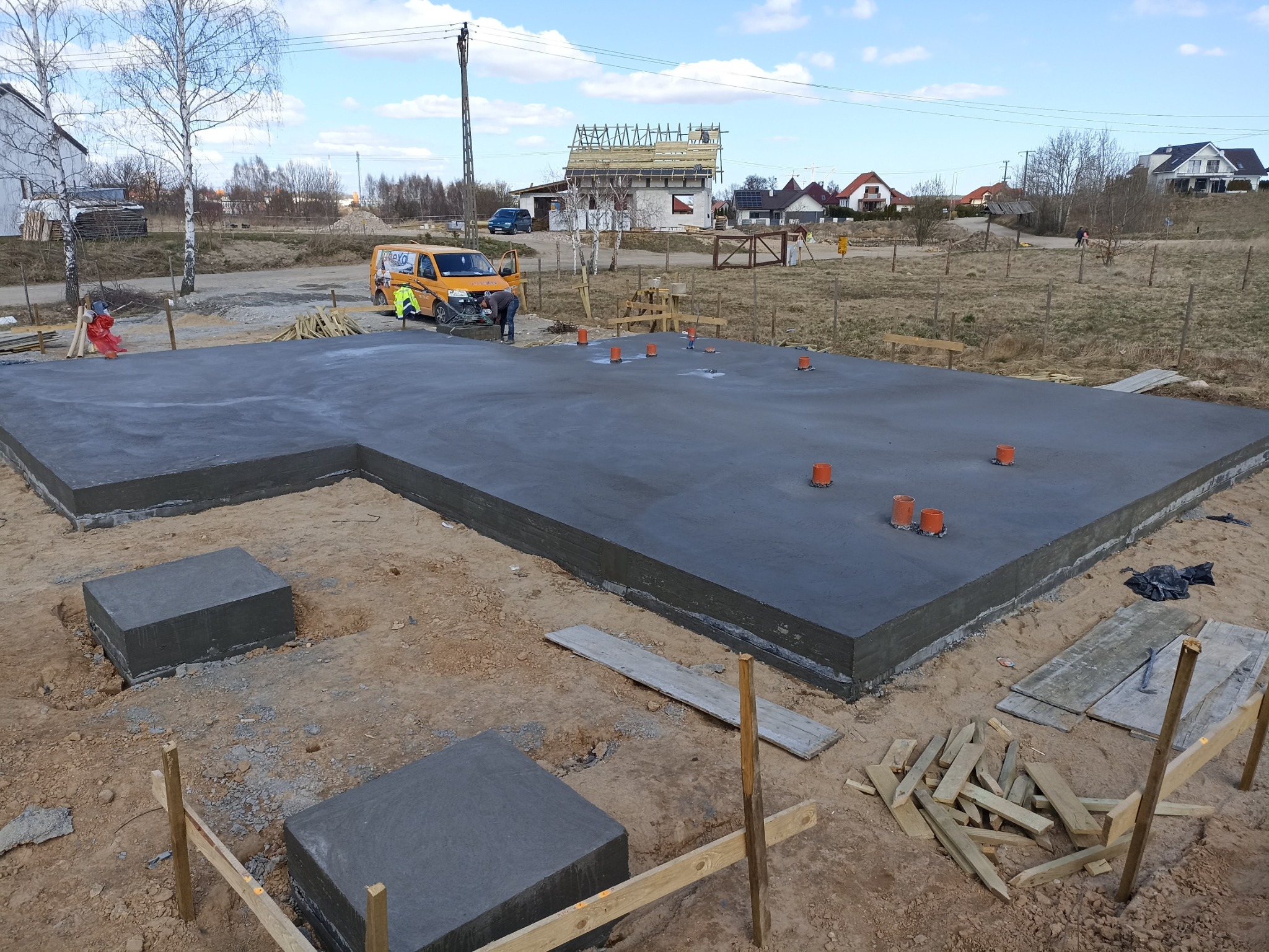 Wylana, gładka płyta fundamentowa z wystającymi rurami instalacyjnymi, widoczny żółty samochód dostawczy i budowa domu w tle.