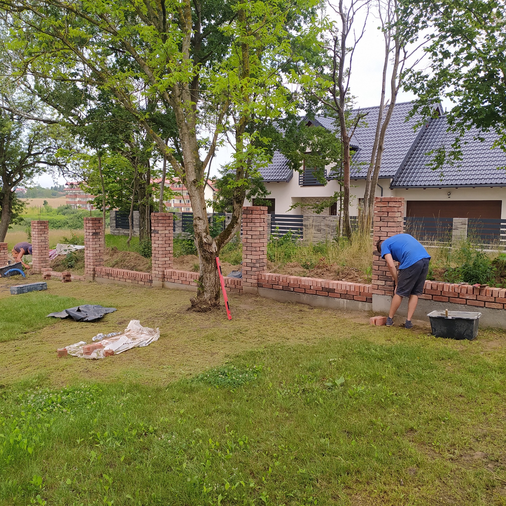 Budowa ogrodzenia z czerwonej cegły, widoczni pracownicy, w tle dom jednorodzinny z ciemnym dachem.