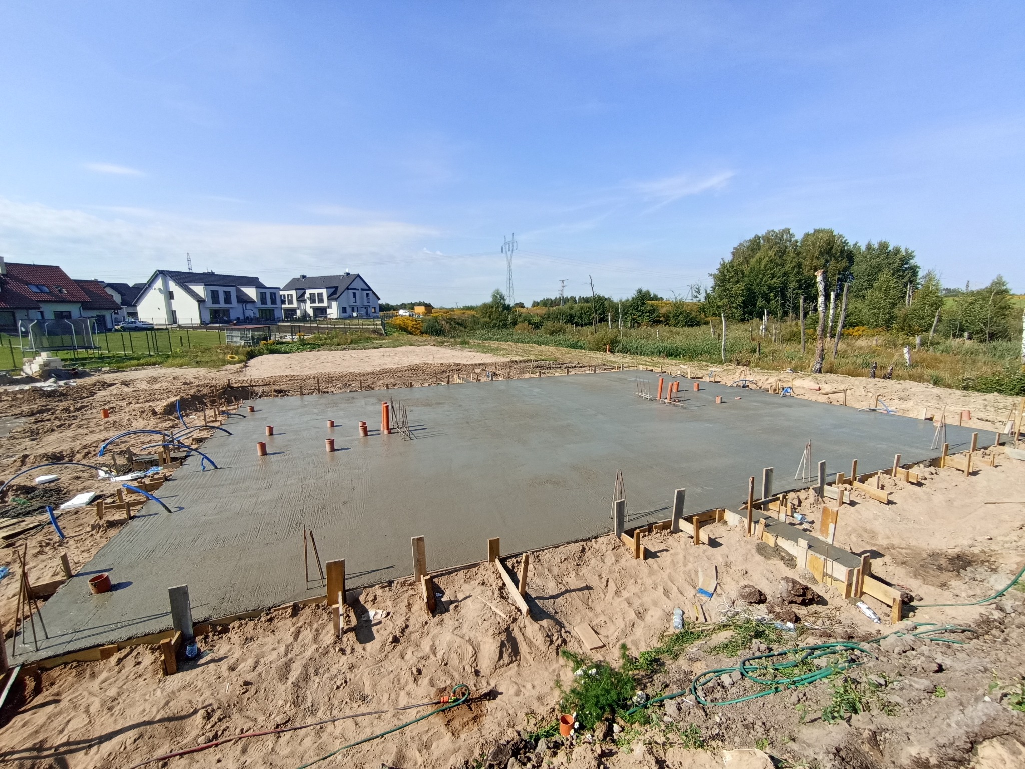 Zalana betonem płyta fundamentowa z wystającymi rurami instalacyjnymi, otoczona drewnianym szalunkiem na piaszczystym terenie budowy. W tle widoczne zabudowania mieszkalne i słupy wysokiego...