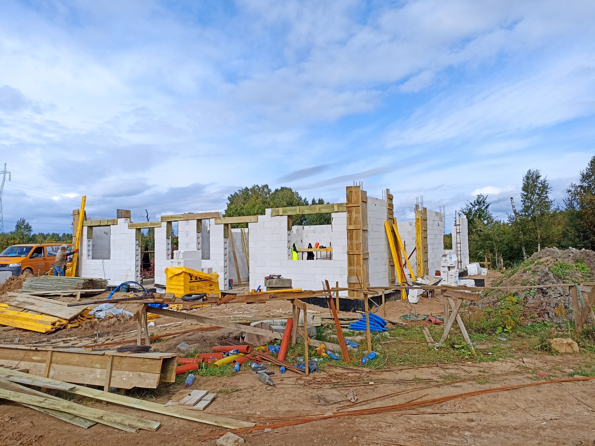 Wznoszenie ścian parterowego domu z białych bloczków, widoczne szalunki i zbrojenia, na pierwszym planie porozrzucane materiały budowlane i narzędzia, w tle częściowo widoczny samochód dostawczy.