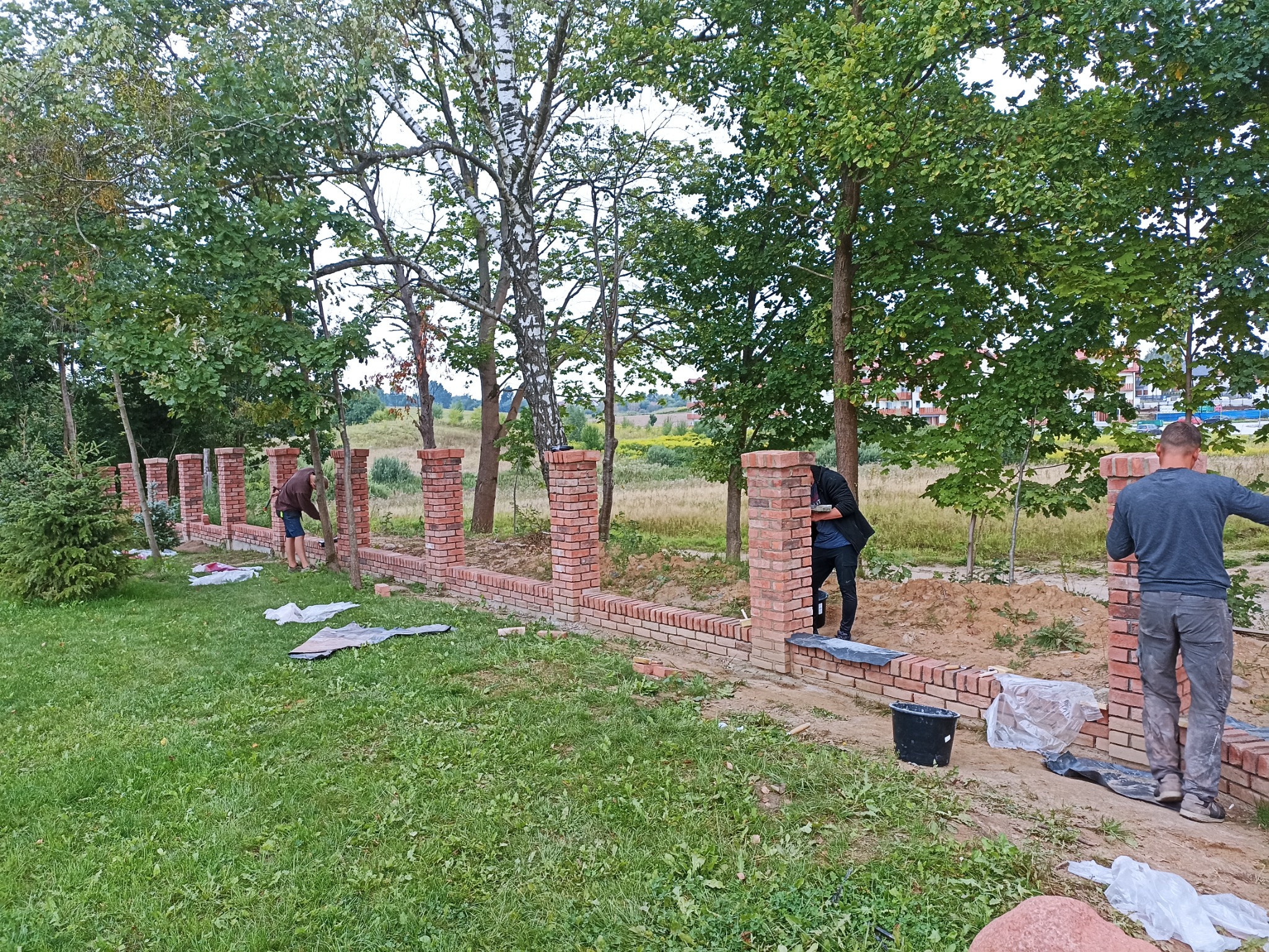 Wznoszenie niskiego muru z czerwonej cegły z filarami na fundamencie w ogrodzie, widoczni pracownicy murujący i przygotowujący zaprawę.
