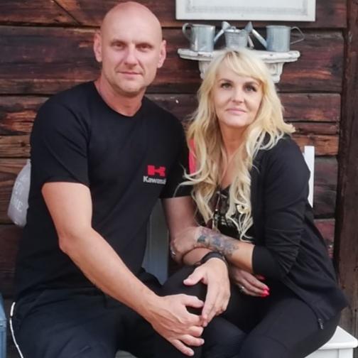 Portret pary siedzącej na ławce przed drewnianą ścianą. Mężczyzna w czarnej koszulce z logo Kawasaki, kobieta z blond włosami i tatuażem na ręce. Na ścianie metalowe konewki.