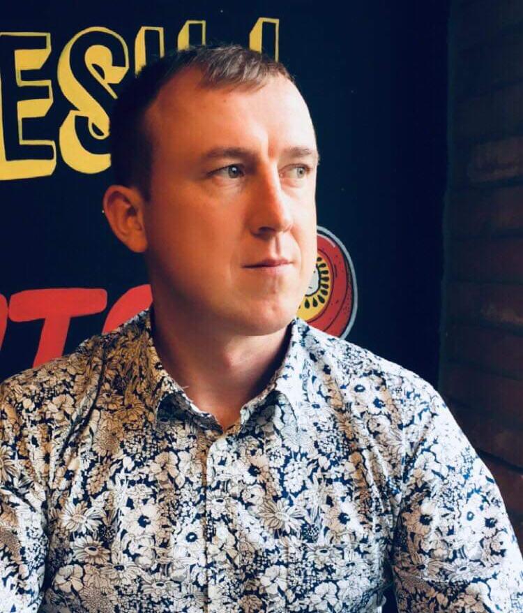 Portret mężczyzny w kwiecistej koszuli na tle ściany z grafiką. Ujęcie z boku, z widocznym fragmentem ceglanej ściany po prawej.