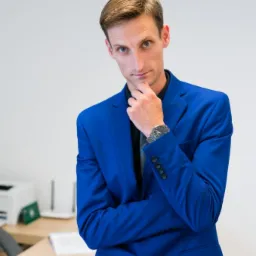 Mężczyzna w niebieskim garniturze, oparty o biurko z dokumentami, w zamyśleniu dotyka brody, spoglądając w obiektyw.