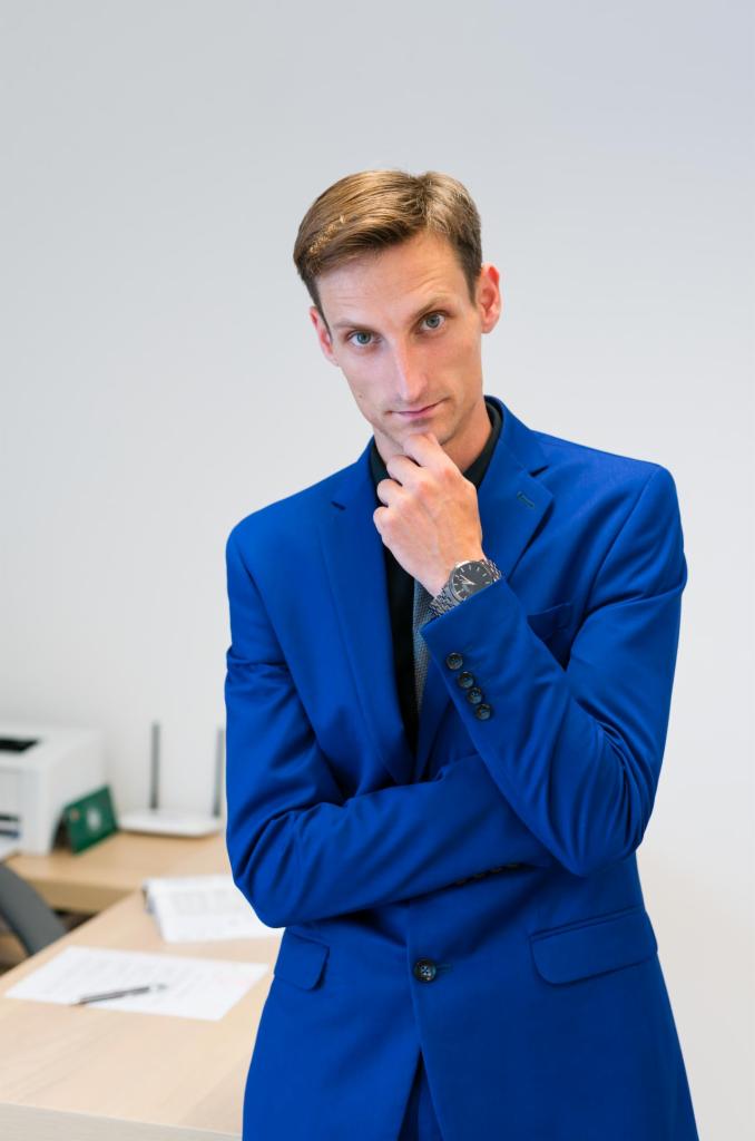 Mężczyzna w niebieskim garniturze, oparty o biurko z dokumentami, w zamyśleniu dotyka brody, spoglądając w obiektyw.