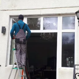 Mężczyzna w szarym kombinezonie stojący na aluminiowej drabinie, montujący białe okno z wieloma szybami w elewacji budynku, widoczny fragment żyrandola wewnątrz.