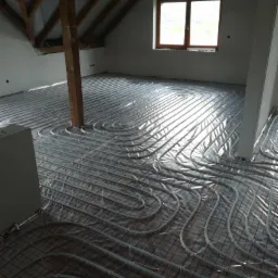 Montaż rur ogrzewania podłogowego na folii izolacyjnej z rastrem w surowym wnętrzu poddasza z drewnianą konstrukcją stropu i oknem.