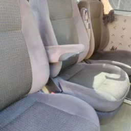 Wnętrze samochodu z widocznymi szarymi fotelami z tapicerką w drobny wzór, przednie fotele z podłokietnikami, widoczne zagłówki i fragmenty tylnych siedzeń.