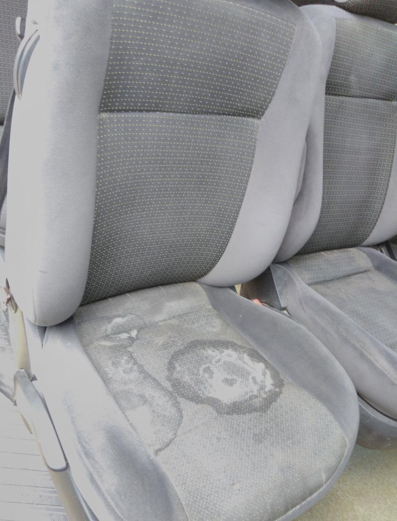 Tapicerka samochodowa z widocznymi plamami i zabrudzeniami na siedzisku, szaro-czarna kolorystyka.