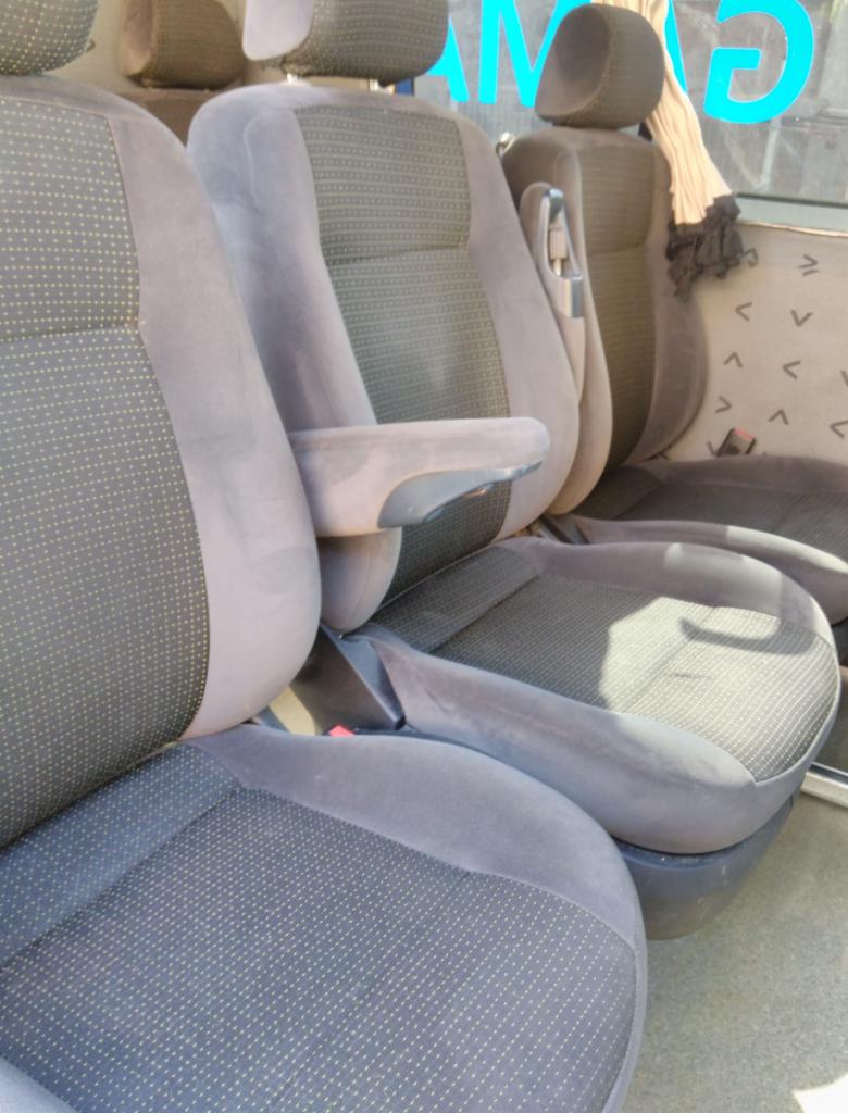 Wnętrze samochodu z widocznymi szarymi fotelami z tapicerką w drobny wzór, przednie fotele z podłokietnikami, widoczne zagłówki i fragmenty tylnych siedzeń.