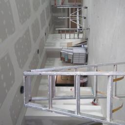 Budullux - Wnętrze w trakcie szpachlowania ścian i sufitu, widoczne ślady po łatach gipsowych, aluminiowa drabina na pierwszym planie, w tle rusztowania i pojemnik z materiałem budowlanym.