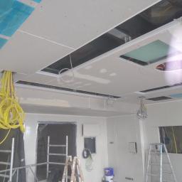 Budullux - Wnętrze w trakcie prac wykończeniowych w Małym Gowinie: widoczne szpachlowane ściany, częściowo zamontowany sufit podwieszany z otworami na oświetlenie, wystające przewody elektryczne i drabiny...