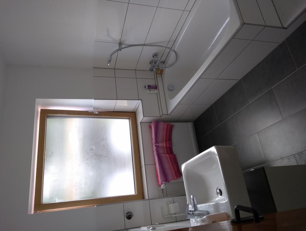 Wykończona łazienka z białymi i szarymi płytkami, drewnianą ramą okienną i wiszącym ręcznikiem w odcieniach różu i fioletu.