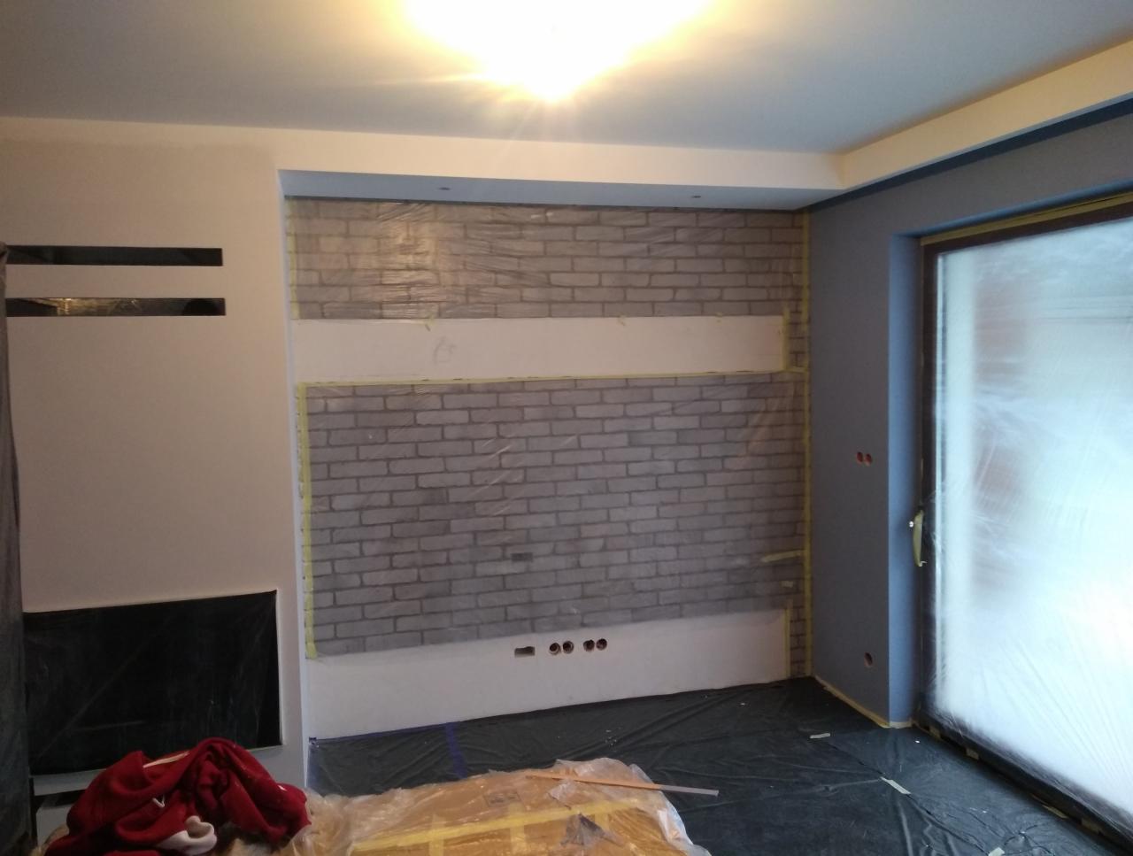 Pokój w trakcie remontu: ściana z cegieł wyklejona taśmą malarską i folią ochronną, przygotowana do dalszych prac wykończeniowych, podłoga zabezpieczona folią, widoczne gniazdka elektryczne.