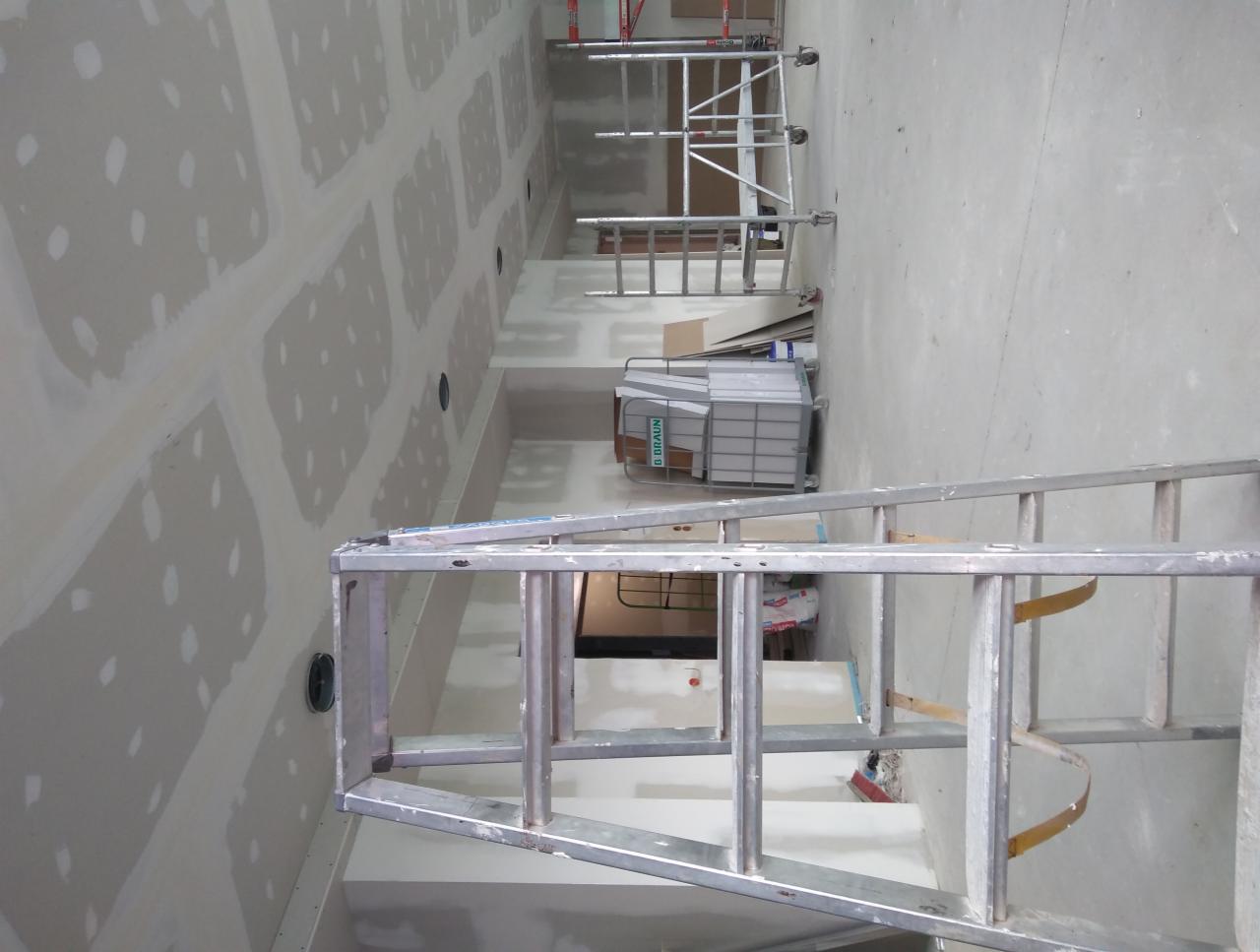 Wnętrze w trakcie szpachlowania ścian i sufitu, widoczne ślady po łatach gipsowych, aluminiowa drabina na pierwszym planie, w tle rusztowania i pojemnik z materiałem budowlanym.