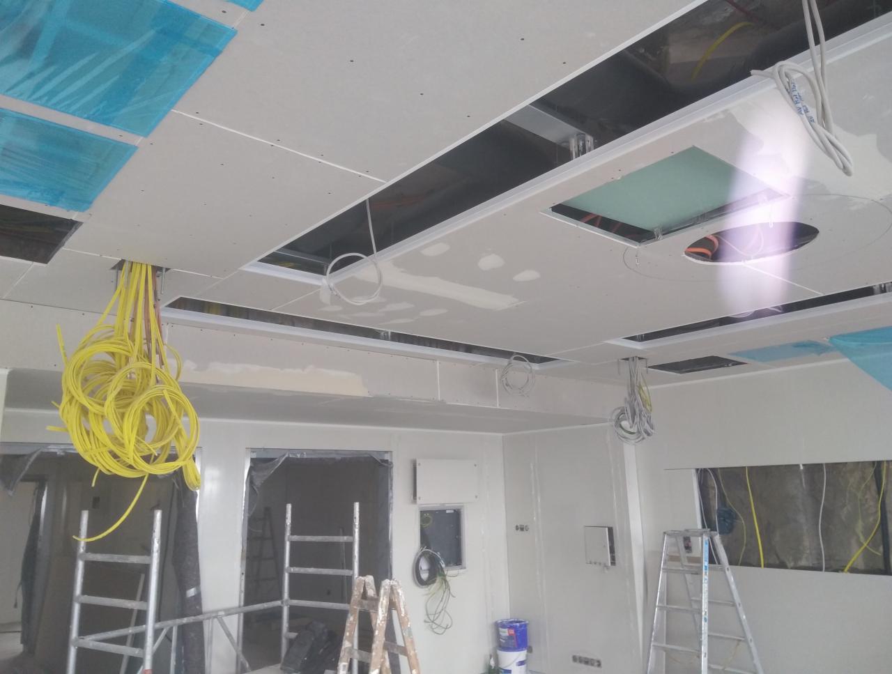 Wnętrze w trakcie prac wykończeniowych w Małym Gowinie: widoczne szpachlowane ściany, częściowo zamontowany sufit podwieszany z otworami na oświetlenie, wystające przewody elektryczne i drabiny...