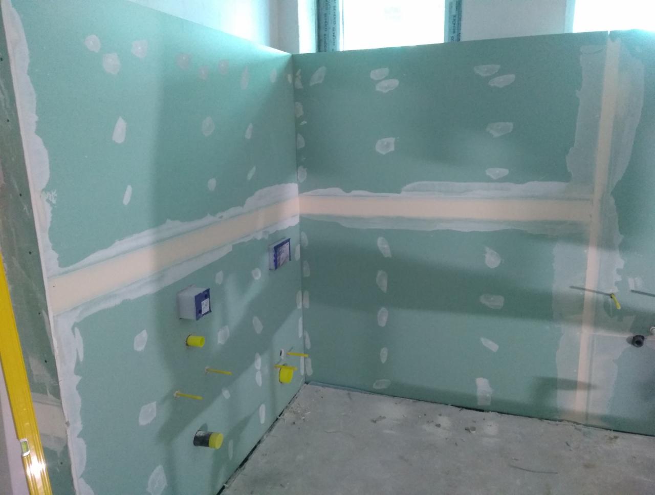 Ściany w narożniku pomieszczenia przygotowane do dalszych prac wykończeniowych, widoczne płyty gipsowo-kartonowe z widocznymi łączeniami i miejscami po szpachlowaniu, wystające elementy instalacji...