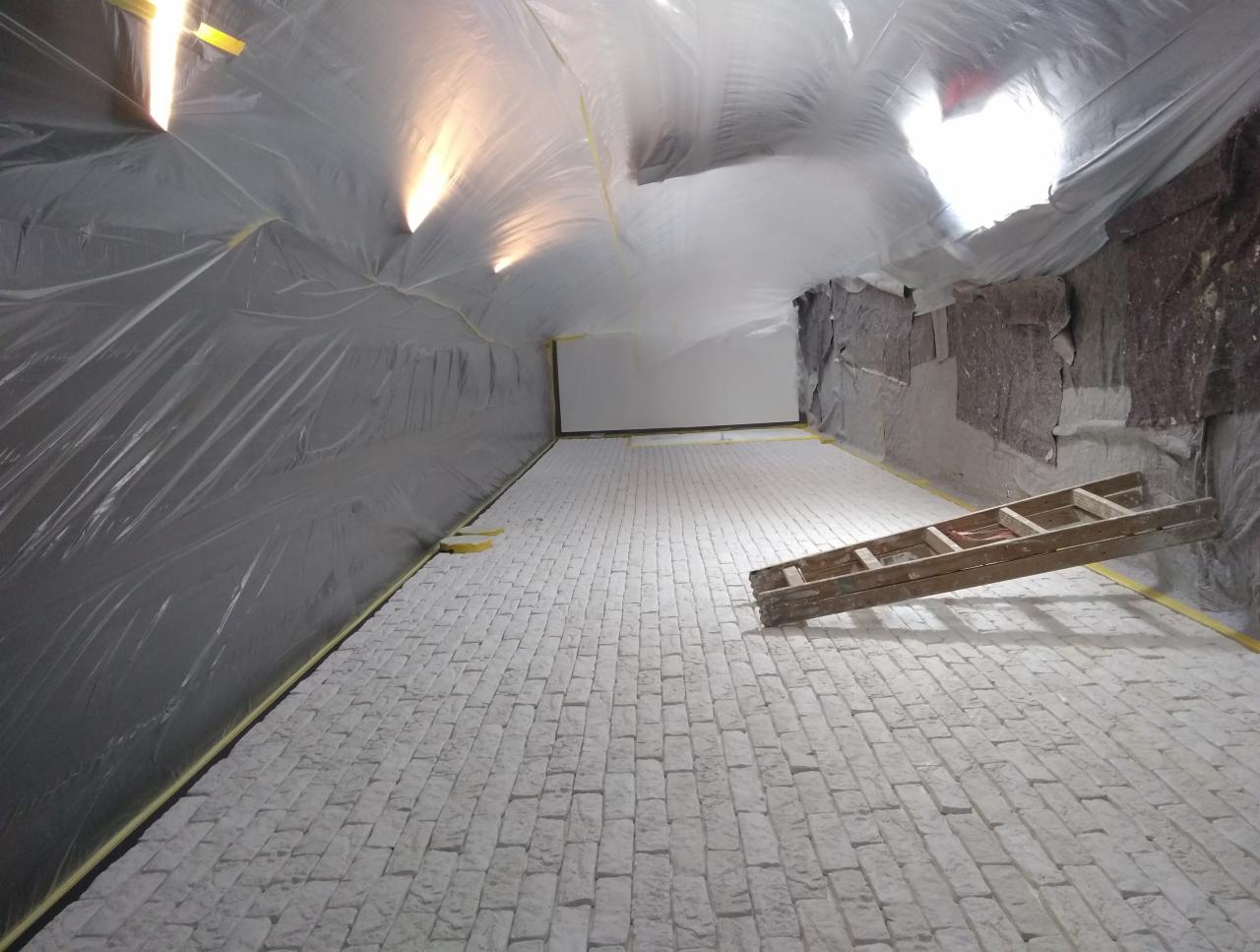 Przygotowane pomieszczenie do prac wykończeniowych z folią ochronną na ścianach i suficie, biała podłoga imitująca cegłę, drewniana drabina oparta o ścianę.