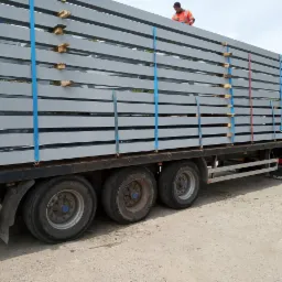 Transport belek stalowych na naczepie ciężarówki, zabezpieczonych niebieskimi pasami, z widocznym pracownikiem w odblaskowej kamizelce na górze ładunku.