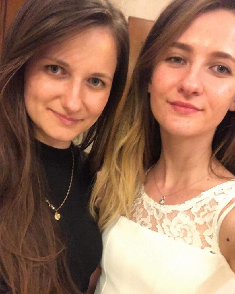 Portret dwóch uśmiechniętych kobiet z długimi włosami, jedna w czarnej bluzce z naszyjnikiem, druga w białej koronkowej sukience. Zdjęcie wewnątrz pomieszczenia.