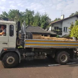 Ciężarówka Volvo FL6 z ładunkiem piasku na pace, kierowana przez mężczyznę. W tle domy i drzewa. Flaga Polski powiewa na pace pojazdu.